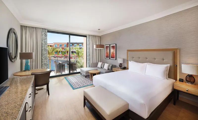Chambre lumineuse avec un grand lit et un espace salon donnant sur une piscine.
