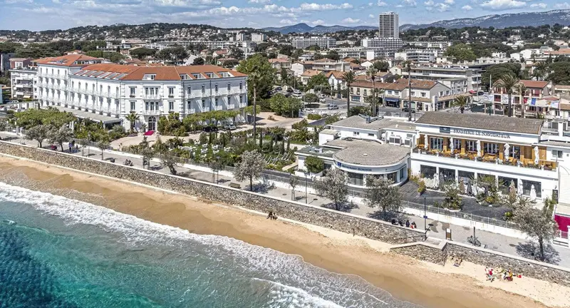 Vue aérienne d'un établissement au bord de mer avec une plage de sable et une ville en arrière-plan.