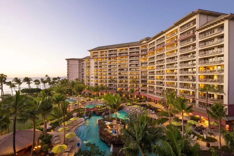 Bâtiment à plusieurs étages avec des balcons illuminés, entouré de végétation tropicale et d'une piscine.
