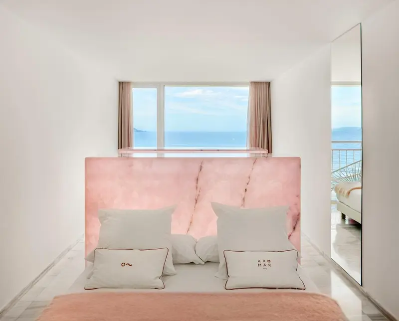Un lit avec une tête de lit rose domine une chambre lumineuse donnant sur la mer.