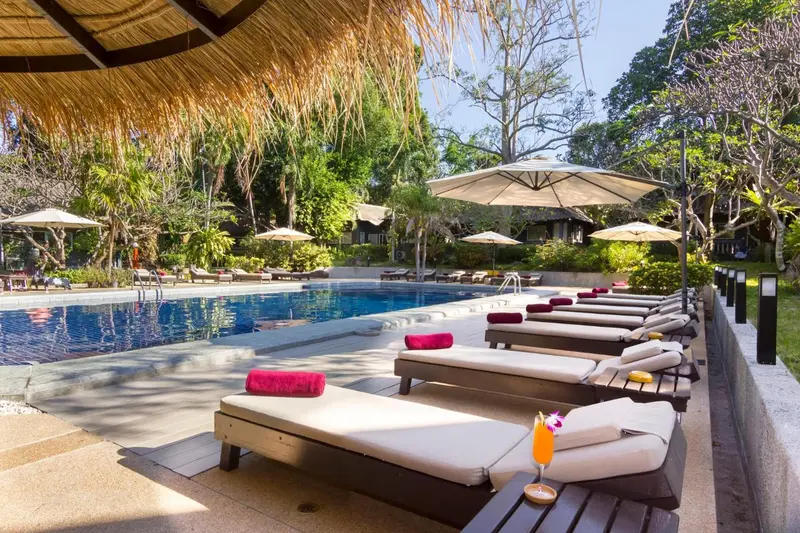 Transats alignés au bord d'une piscine ensoleillée, avec parasols et végétation tropicale.