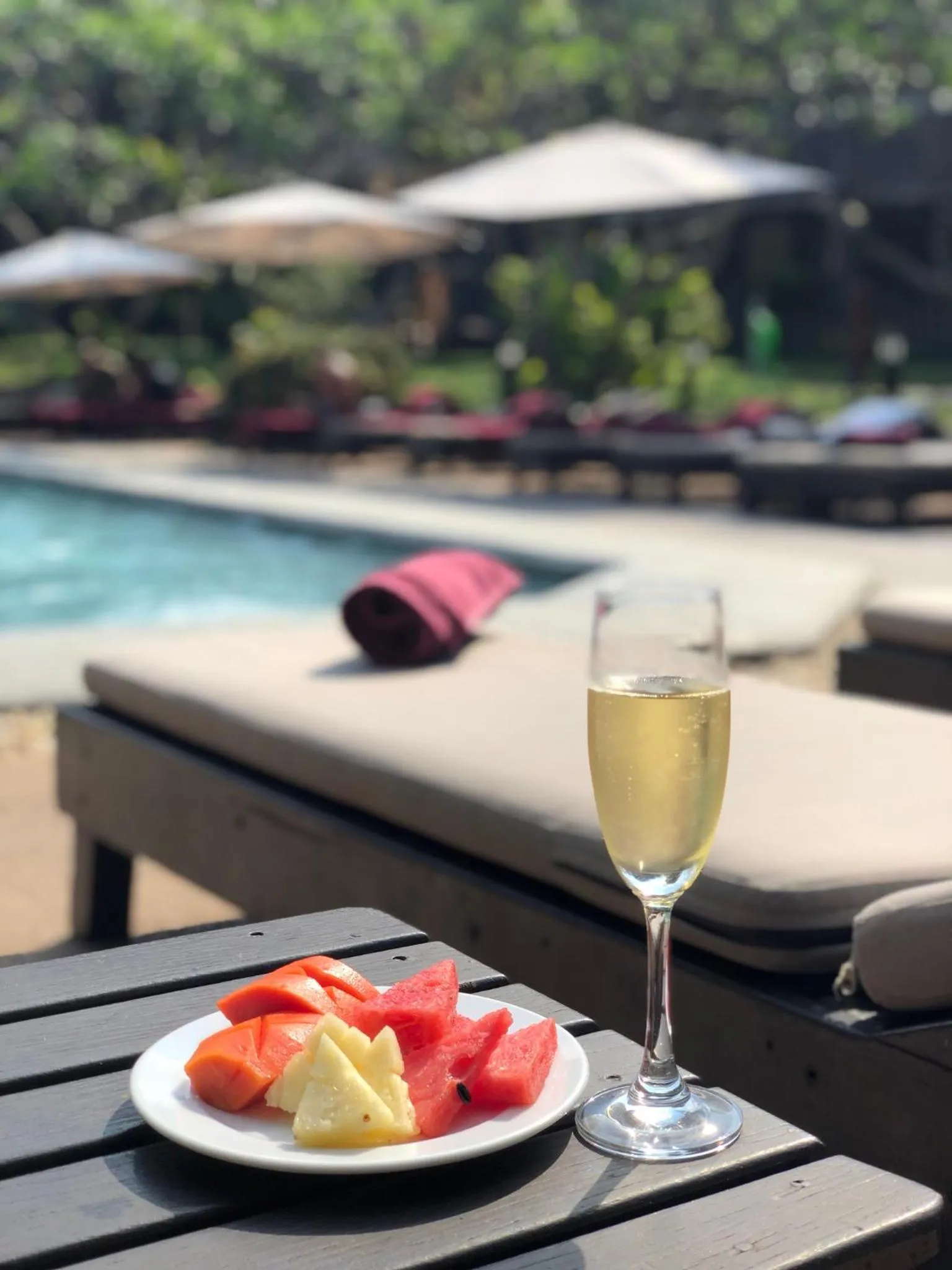 Une table basse en bois présente une assiette de fruits frais et un verre de boisson, près d'une piscine avec des transats.