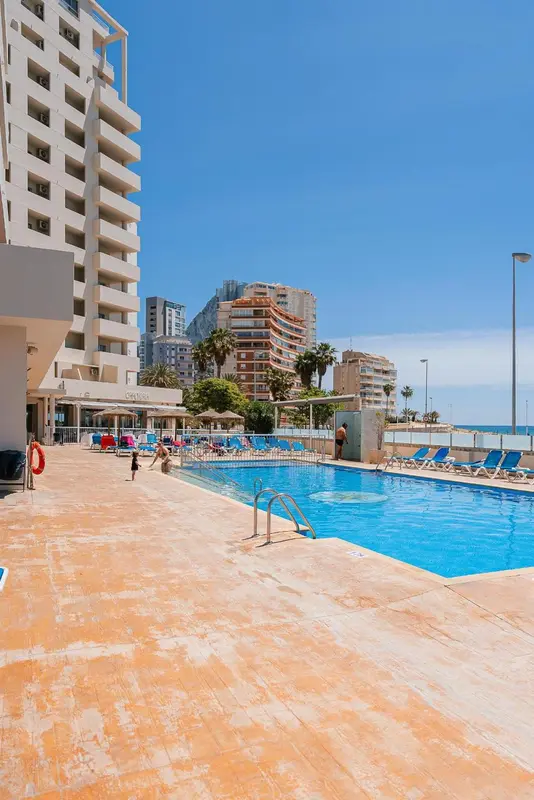 Une piscine turquoise bordée d'une terrasse ensoleillée devant un bâtiment moderne.