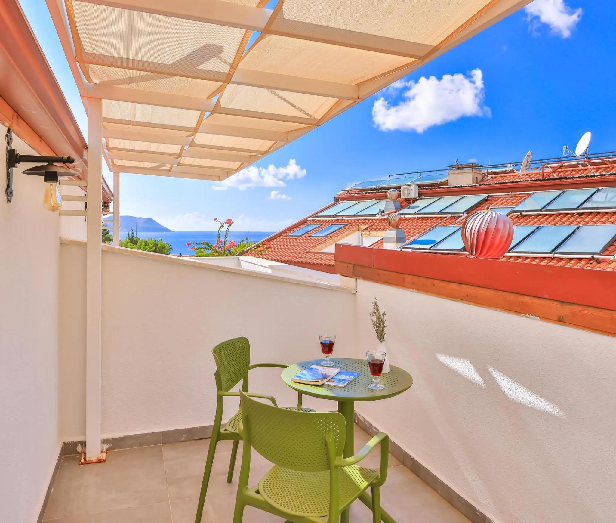 Une table ronde et deux chaises vertes se trouvent sur une terrasse ombragée avec vue sur la mer.