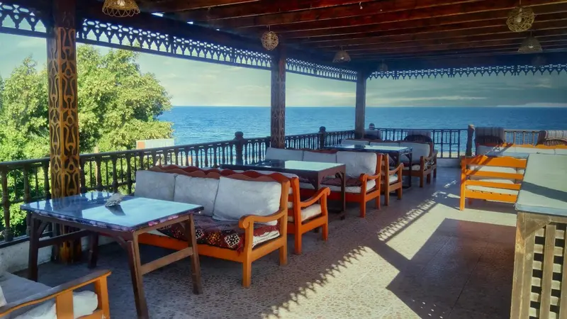 Terrasse ombragée avec tables et canapés donnant sur la mer.