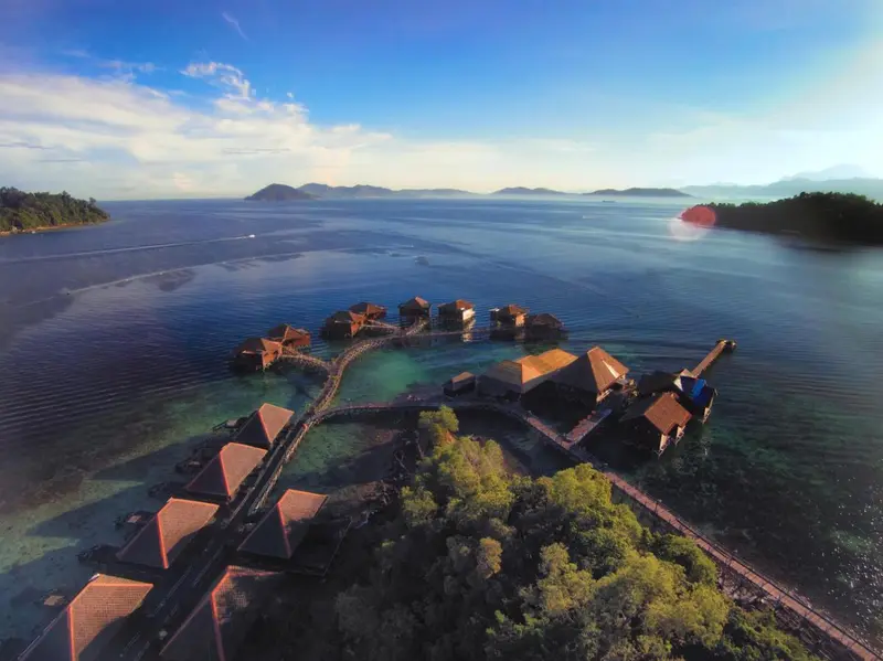 Vue aérienne d'un ensemble de bungalows sur pilotis au-dessus d'une eau turquoise.