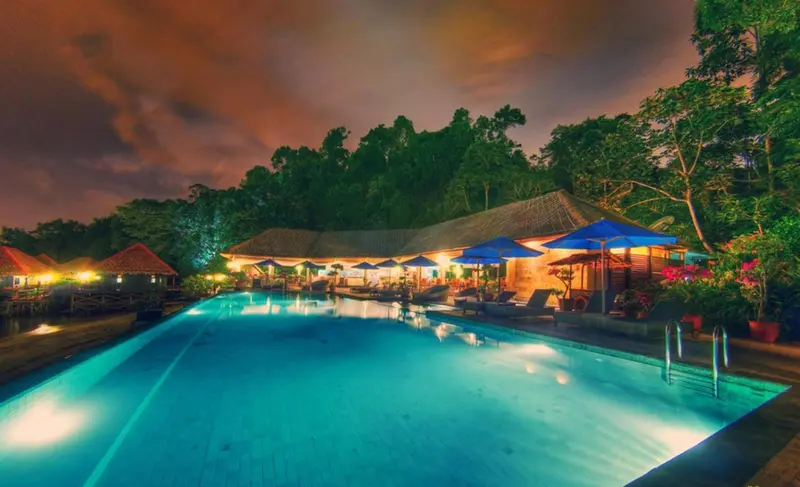 Piscine illuminée au crépuscule devant un bâtiment tropical.