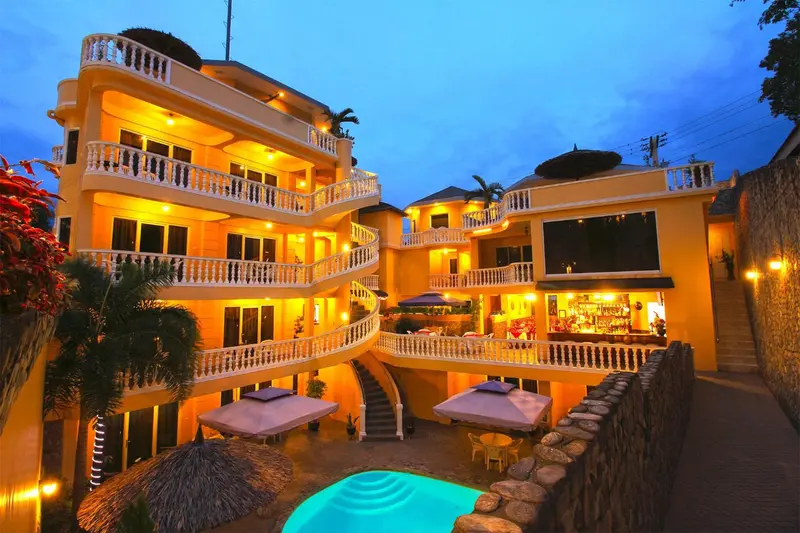 Bâtiment jaune à plusieurs étages illuminé la nuit avec une piscine en contrebas.