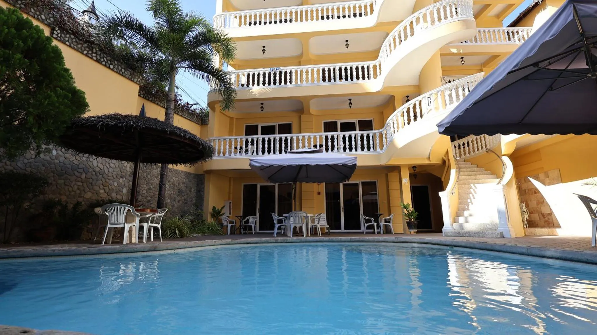 Bâtiment jaune à balcons surplombant une piscine et une terrasse ombragée.