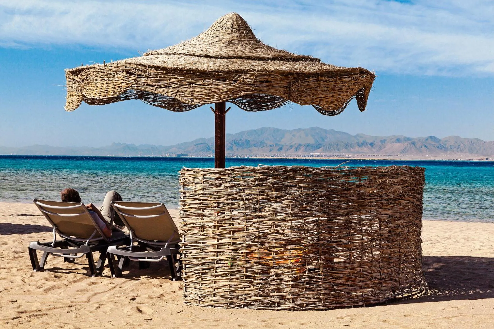 Deux transats et un parasol en osier sur une plage de sable fin avec vue sur la mer et les montagnes.