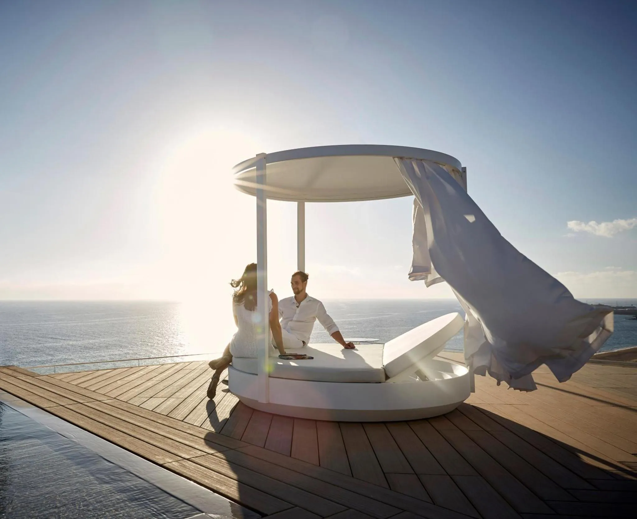 Un couple se détend sur un lit de jour rond au bord d'une piscine à débordement avec vue sur la mer.