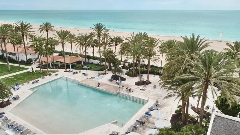 Vue aérienne d'une piscine bordée de transats, avec une plage et la mer en arrière-plan.