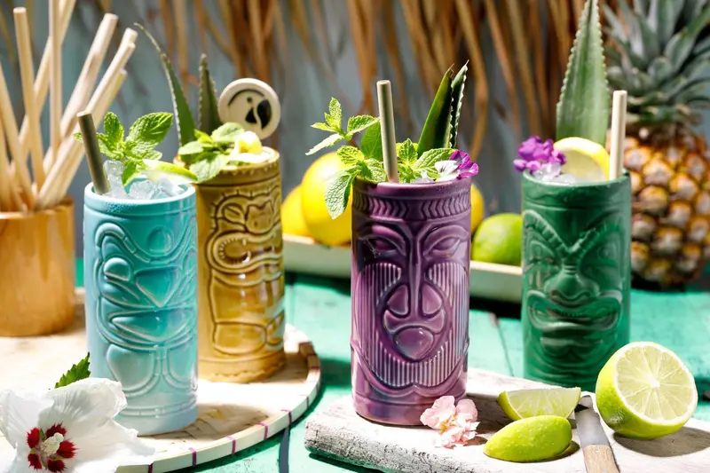 Quatre cocktails colorés servis dans des verres tiki décorés de fruits et de fleurs.