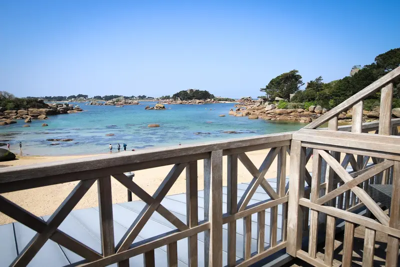Vue sur une plage de sable fin et une mer turquoise depuis une terrasse en bois.
