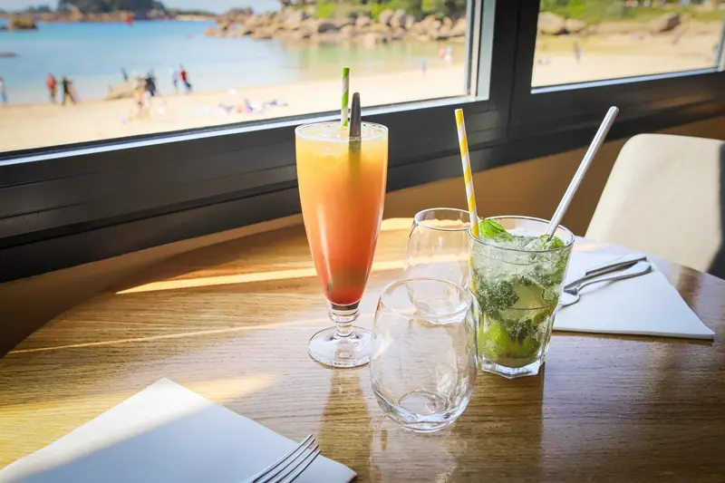 Deux cocktails colorés et des verres sur une table en bois, avec une vue sur une plage ensoleillée.