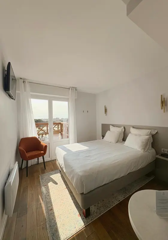 Une chambre lumineuse avec un lit fait et un fauteuil près d'une fenêtre.