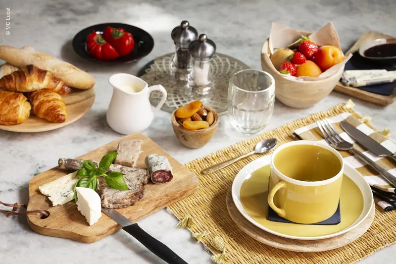 Une table est dressée avec des viennoiseries, des fruits, de la charcuterie et des boissons.