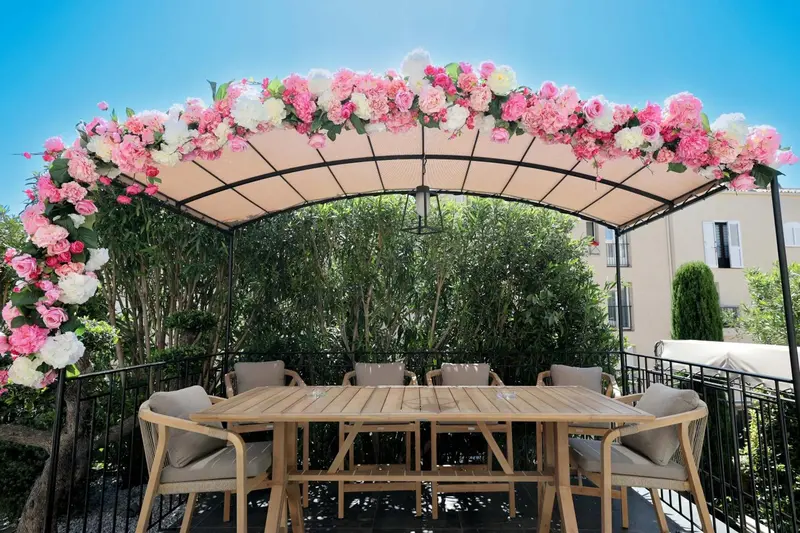 Une table à manger en bois est placée sous une tonnelle fleurie sur une terrasse.