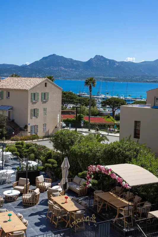 Terrasse ombragée avec tables et chaises donnant sur un port de plaisance et des montagnes.