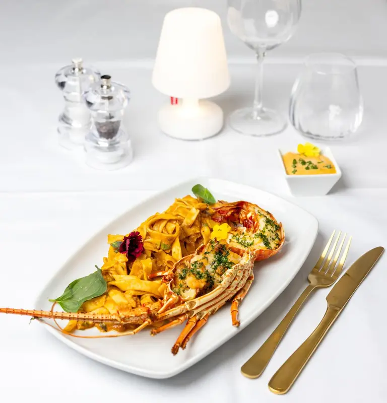 Une assiette de pâtes au homard est présentée avec une fourchette et un couteau dorés sur une table blanche.
