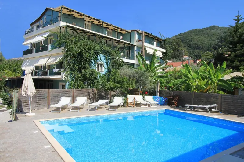 Bâtiment blanc et bleu avec des balcons, entouré de végétation et surplombant une piscine.