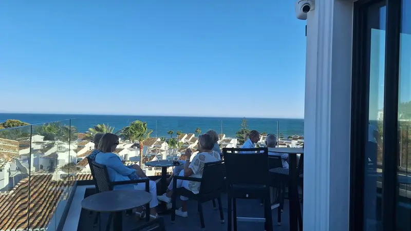Des personnes sont assises à des tables sur une terrasse ensoleillée avec vue sur la mer.