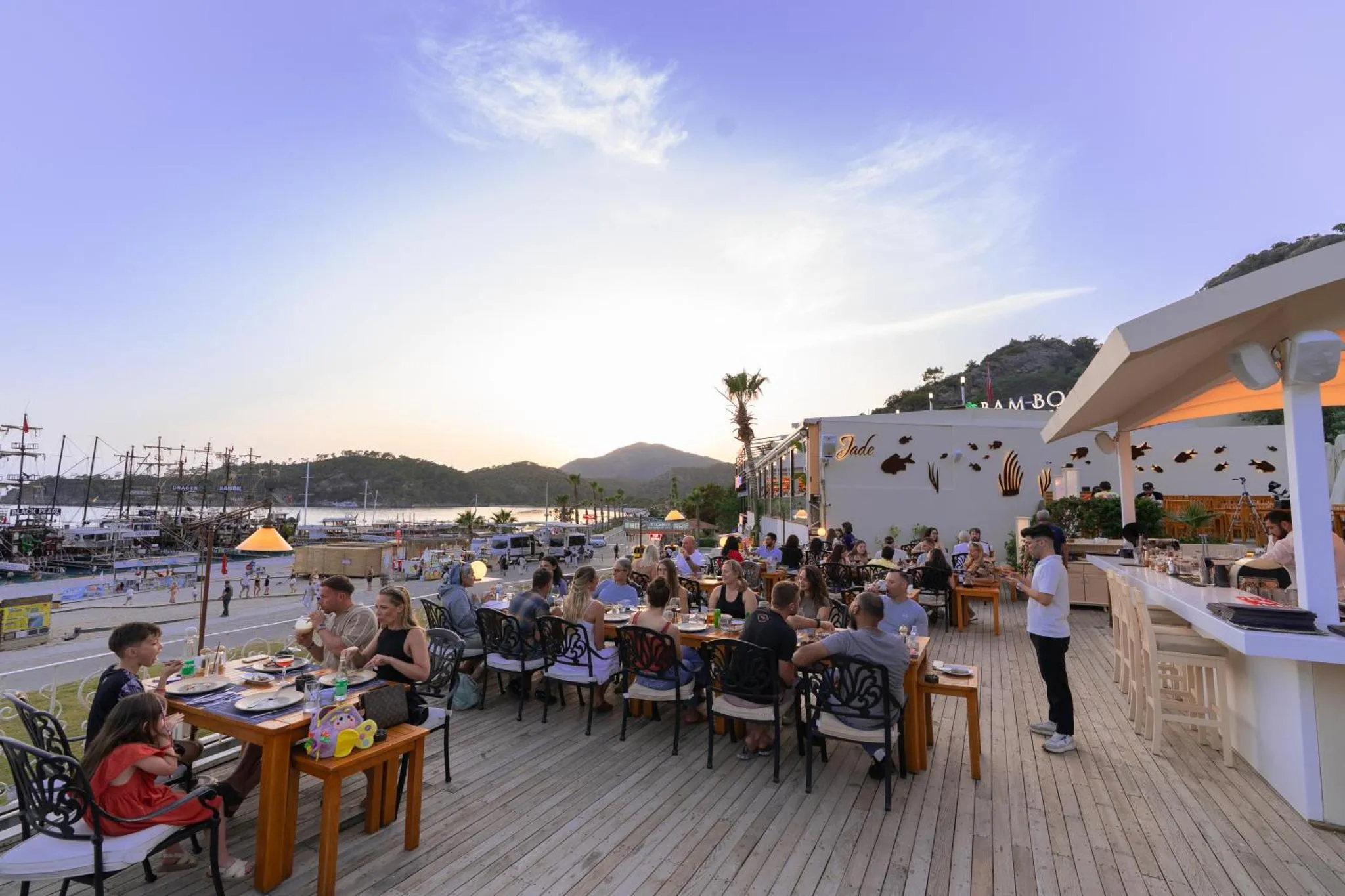 Une terrasse de restaurant animée avec des clients attablés face à la mer au coucher du soleil.