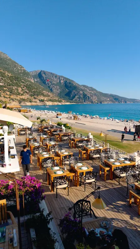 Terrasse de restaurant avec tables dressées face à une plage et des montagnes.