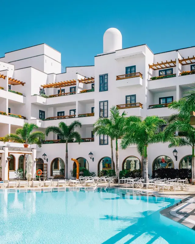 Bâtiment blanc à plusieurs étages avec des balcons et des palmiers, surplombant une piscine turquoise.