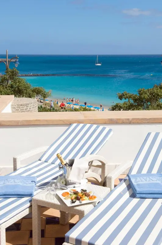 Deux transats rayés avec une bouteille et des fruits sur une terrasse ensoleillée, vue sur la mer.