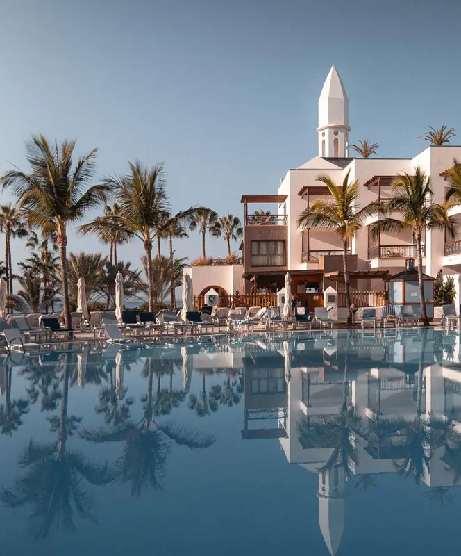 Vue d'une piscine turquoise reflétant un bâtiment blanc et des palmiers.