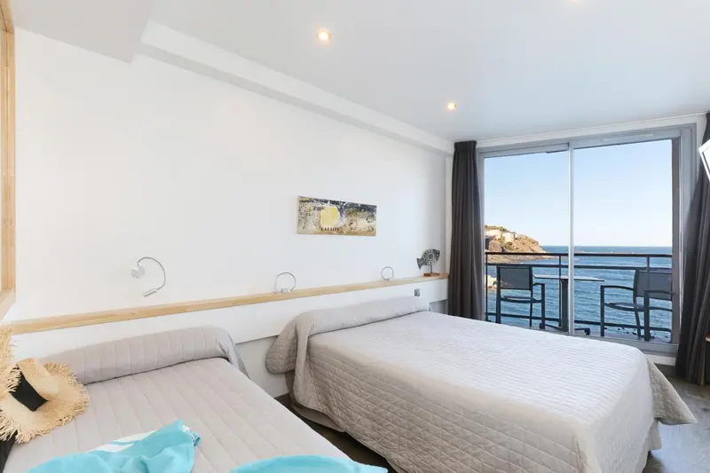 Chambre lumineuse avec deux lits et vue sur la mer depuis une baie vitrée.