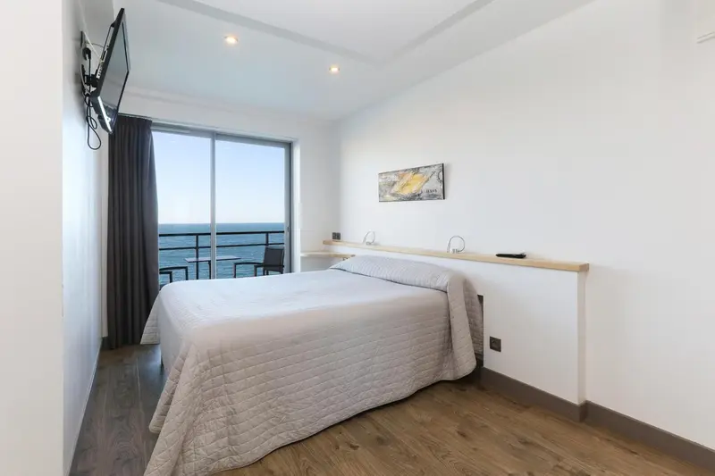 Une chambre lumineuse avec un grand lit face à une baie vitrée donnant sur la mer.