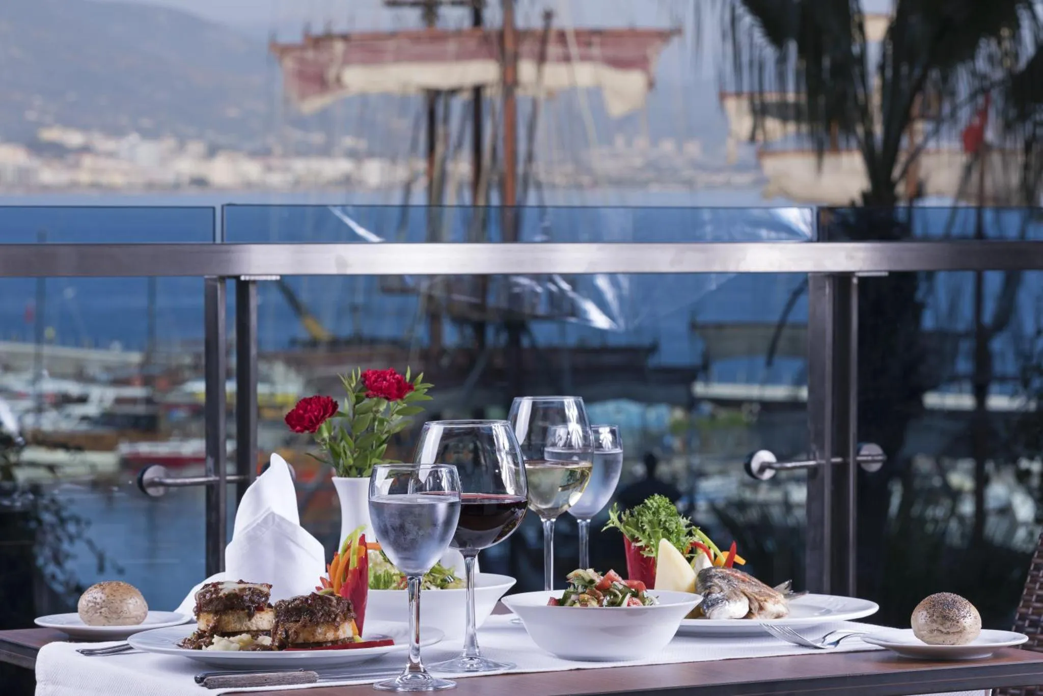 Une table est dressée avec des plats et des boissons, face à une vue sur la mer et un port.