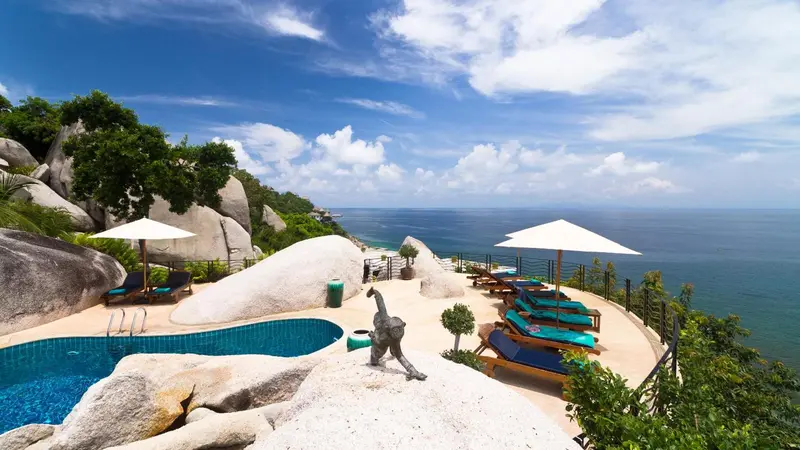 Piscine à débordement avec transats et parasols, surplombant la mer et les rochers.