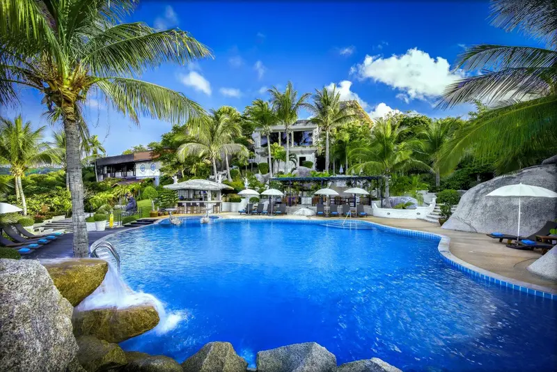 Une piscine turquoise bordée de rochers et de palmiers, avec des transats et un bâtiment en arrière-plan.