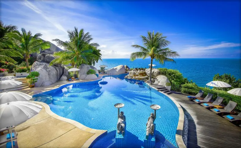 Une piscine bleue bordée de transats et de parasols surplombe la mer.