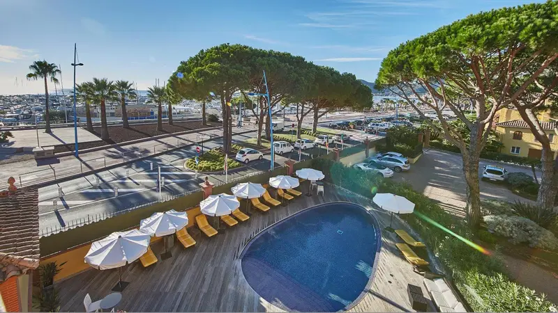 Vue aérienne d'une piscine avec transats et parasols, bordée d'une promenade et d'arbres.