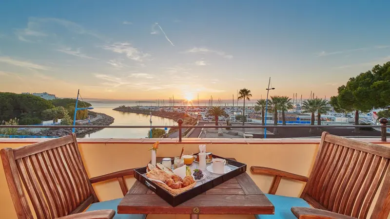 Une table de petit-déjeuner est dressée sur une terrasse avec vue sur un port au lever du soleil.