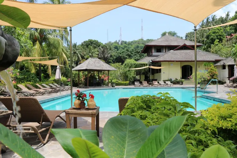 Une piscine turquoise bordée de transats et de végétation luxuriante, avec un bâtiment à l'arrière-plan.