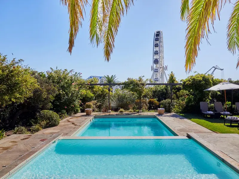 Une piscine rectangulaire bordée d'une terrasse ensoleillée, avec une végétation luxuriante et une grande roue en arrière-plan.