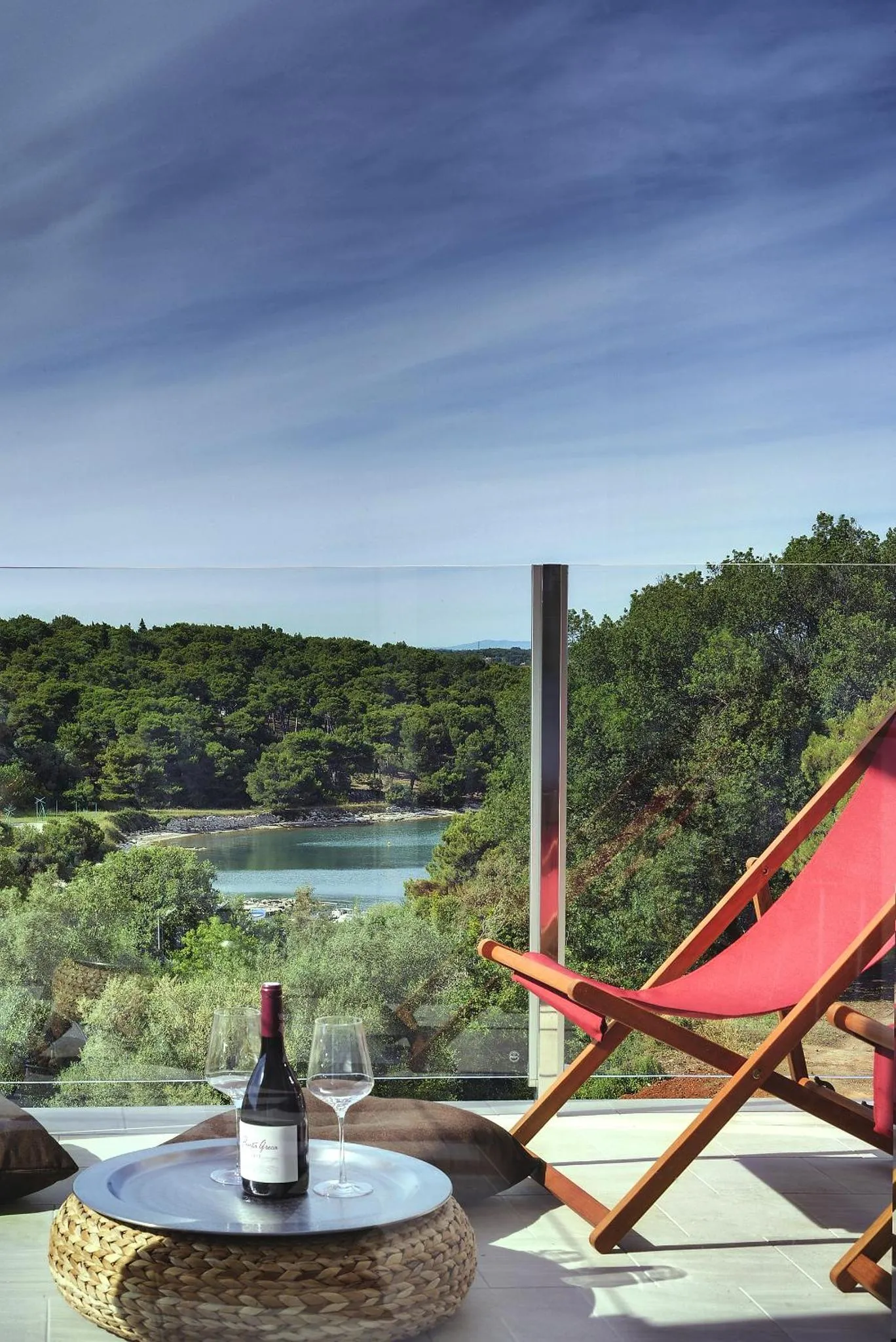 Une table basse avec une bouteille de vin et deux verres sur une terrasse avec vue sur la mer et la végétation.