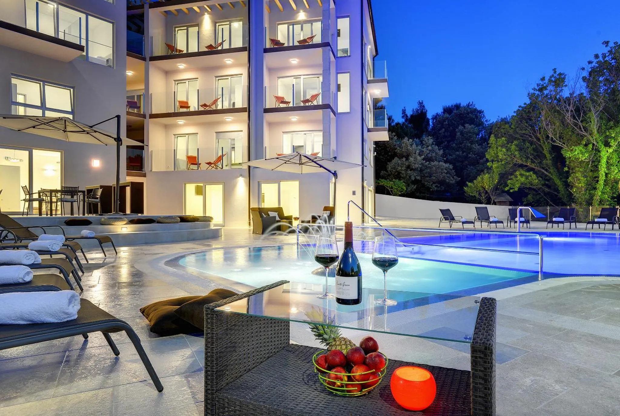 Une table basse avec des boissons et des fruits près d'une piscine éclairée au crépuscule.