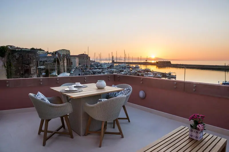 Une table et des fauteuils en osier sont disposés sur une terrasse avec vue sur un port au coucher du soleil.