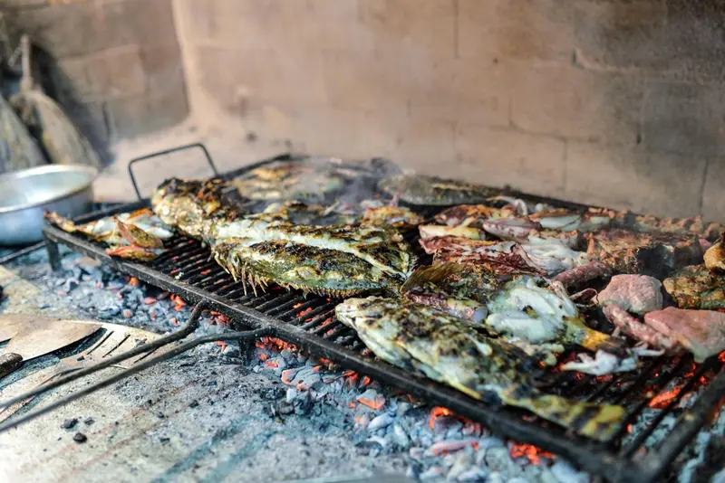 Des poissons et de la viande grillent sur un barbecue en métal au-dessus de braises incandescentes.
