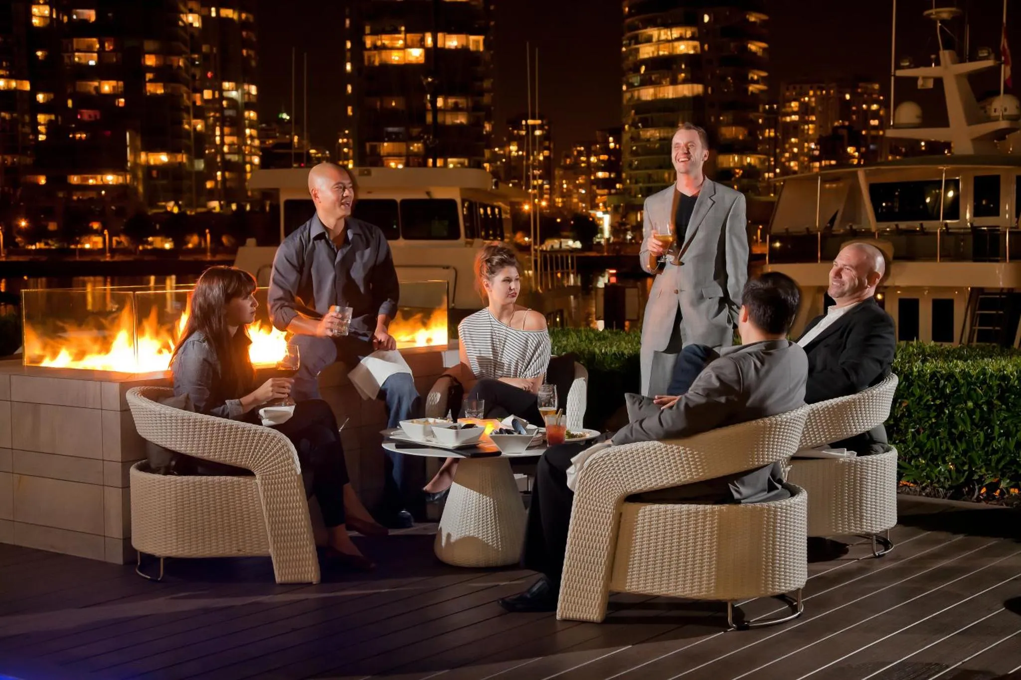 Un groupe de personnes est assis autour d'un foyer extérieur sur une terrasse en soirée.