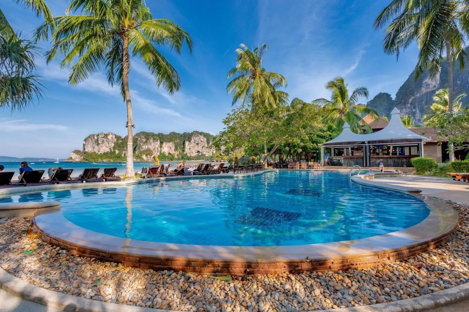 Une piscine turquoise bordée de transats, avec des palmiers et une plage en arrière-plan.