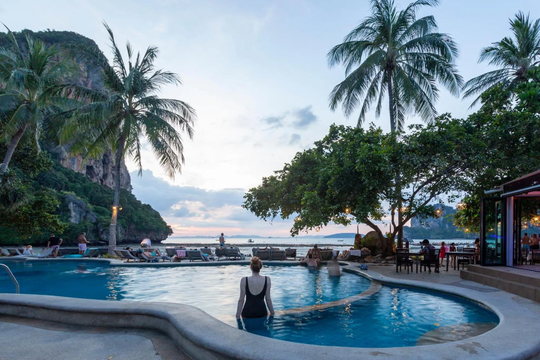 Une piscine bordée de transats, avec des palmiers et une vue sur la mer au loin.