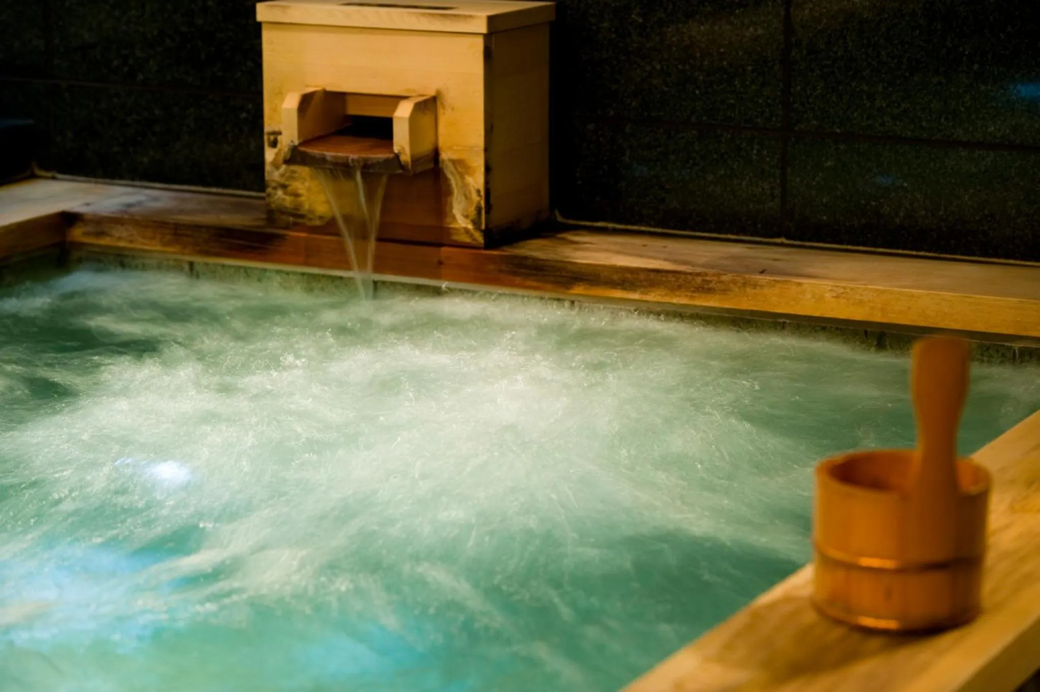 Un bain à remous en bois avec de l'eau turquoise et un seau en bois à proximité.