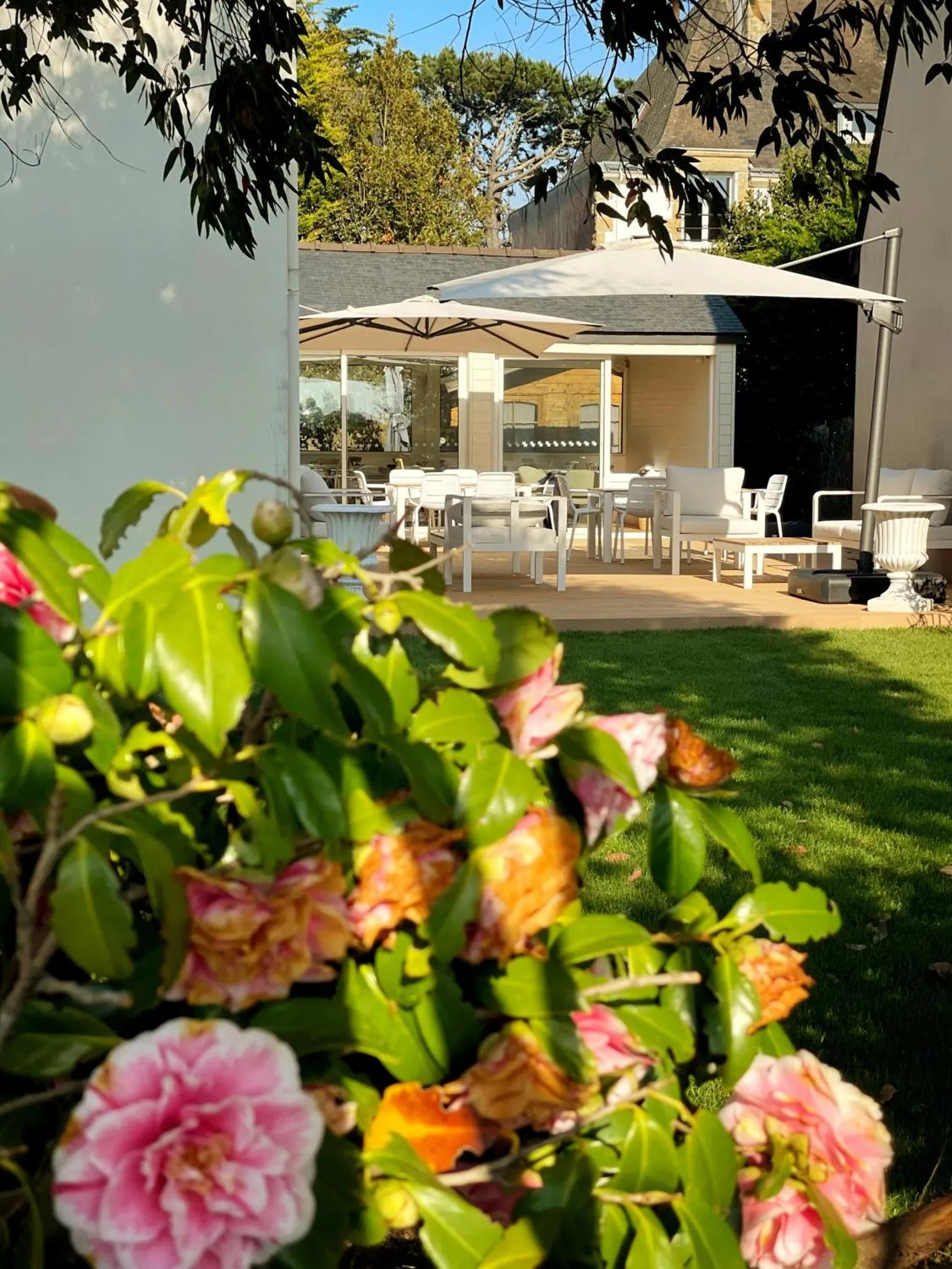 Terrasse ombragée avec tables et chaises blanches, entourée de verdure et de fleurs.
